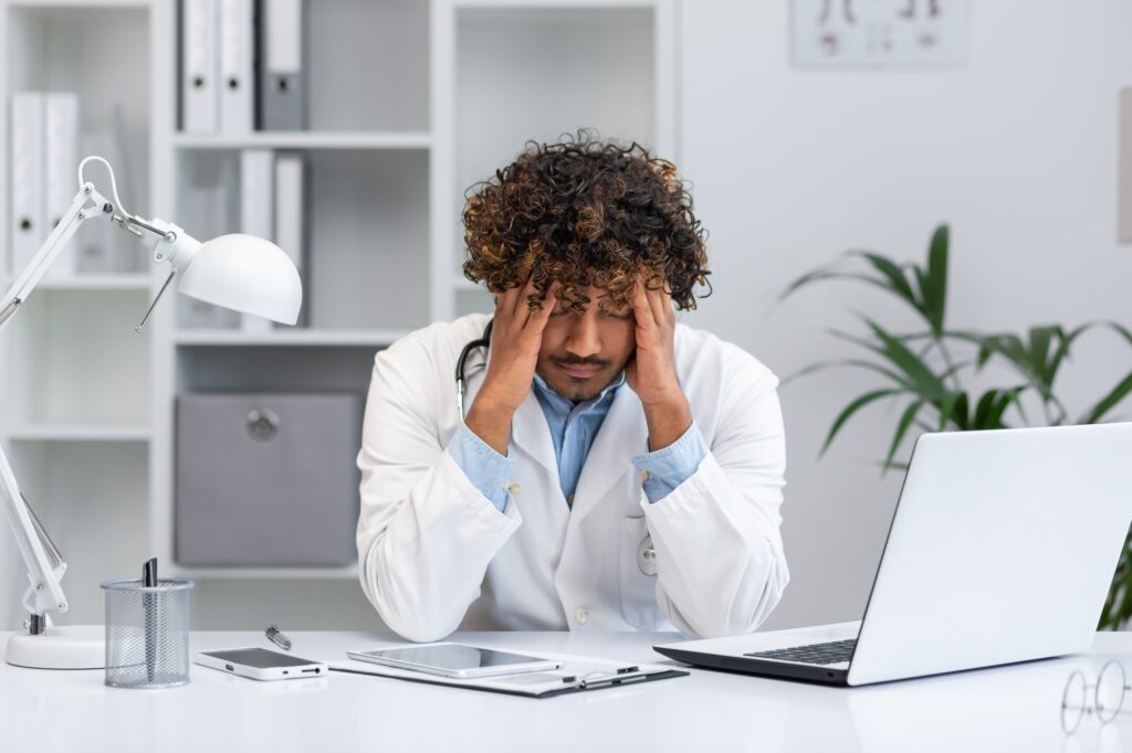 Tired Indian doctor working in medical office, man holding hands on head from overload, sitting at